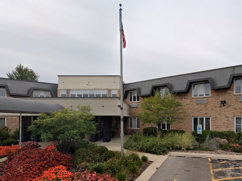 Exterior view of a senior living facility named Wynscape, showing a two-story brick building with a covered entrance, landscaped garden with various shrubs and flowers, a flagpole with an American flag, and a parking area with a handicap parking sign.