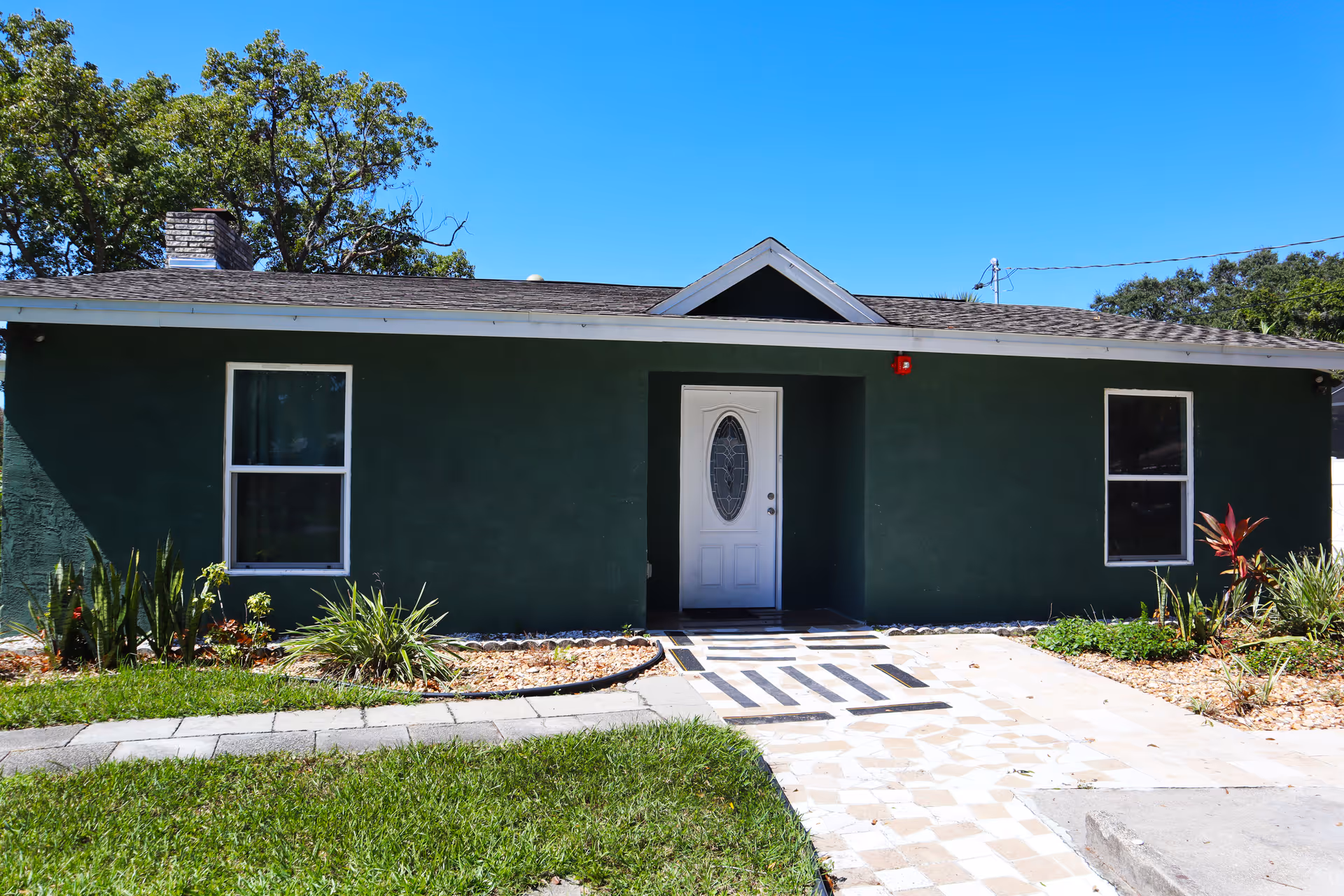 Front exterior view of a single-story green building with a white door and two windows, surrounded by a small garden with plants and a paved walkway leading to the entrance under a clear blue sky.