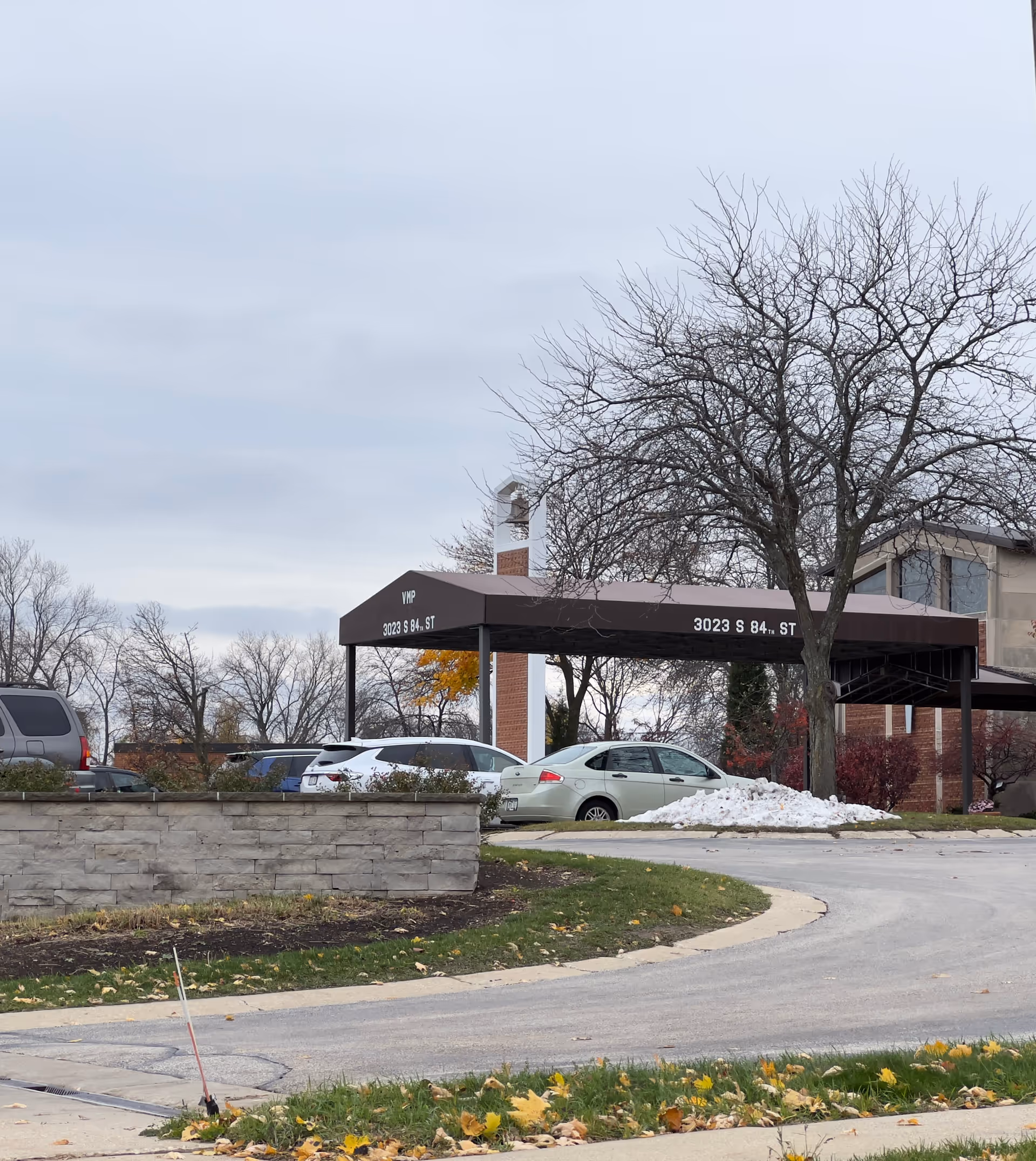 Covered driveway and entrance canopy with address '3023 S 84th St', parked cars, a leafless tree, and a low stone wall in front of a senior living building.