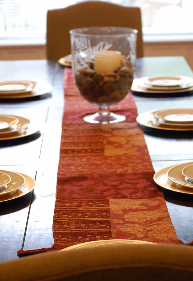 A dining table set with a red and orange patterned table runner, six place settings with gold-rimmed plates, and a centerpiece consisting of a glass container filled with decorative stones and a candle inside.
