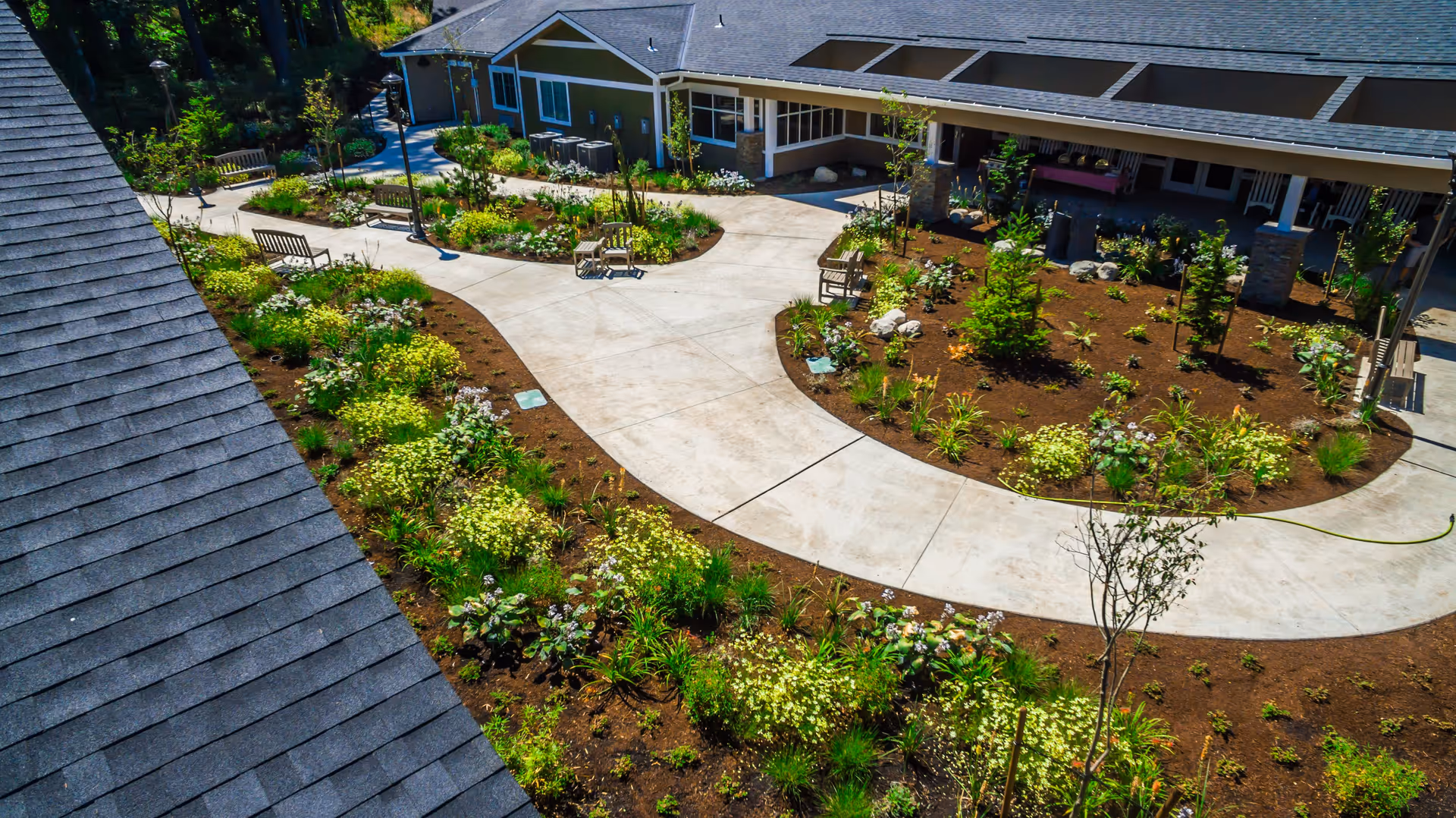 A landscaped outdoor garden area with curved concrete walkways, benches, and various plants and shrubs surrounding a single-story building with a covered porch.