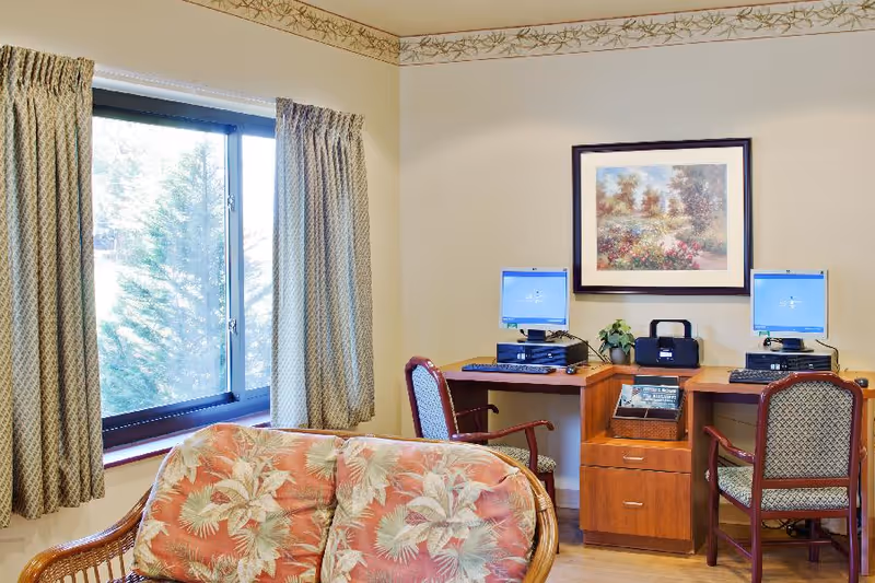 Common sitting area with a floral-upholstered couch, two computer stations on a wooden desk, a framed painting, and a curtained window.