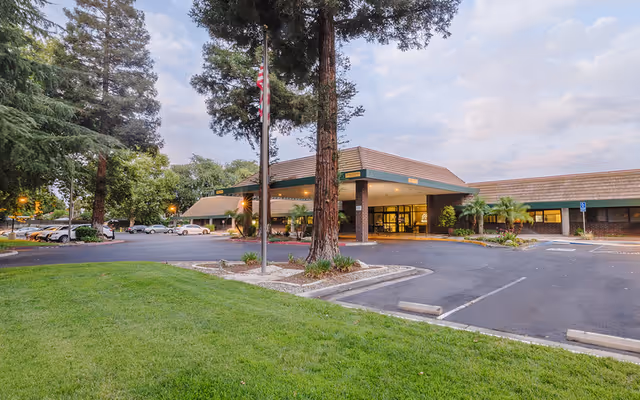 Front entrance of Willow Creek Healthcare Center with a covered drop-off, flagpole, parking spaces, and large trees on a grassy lot at dusk.