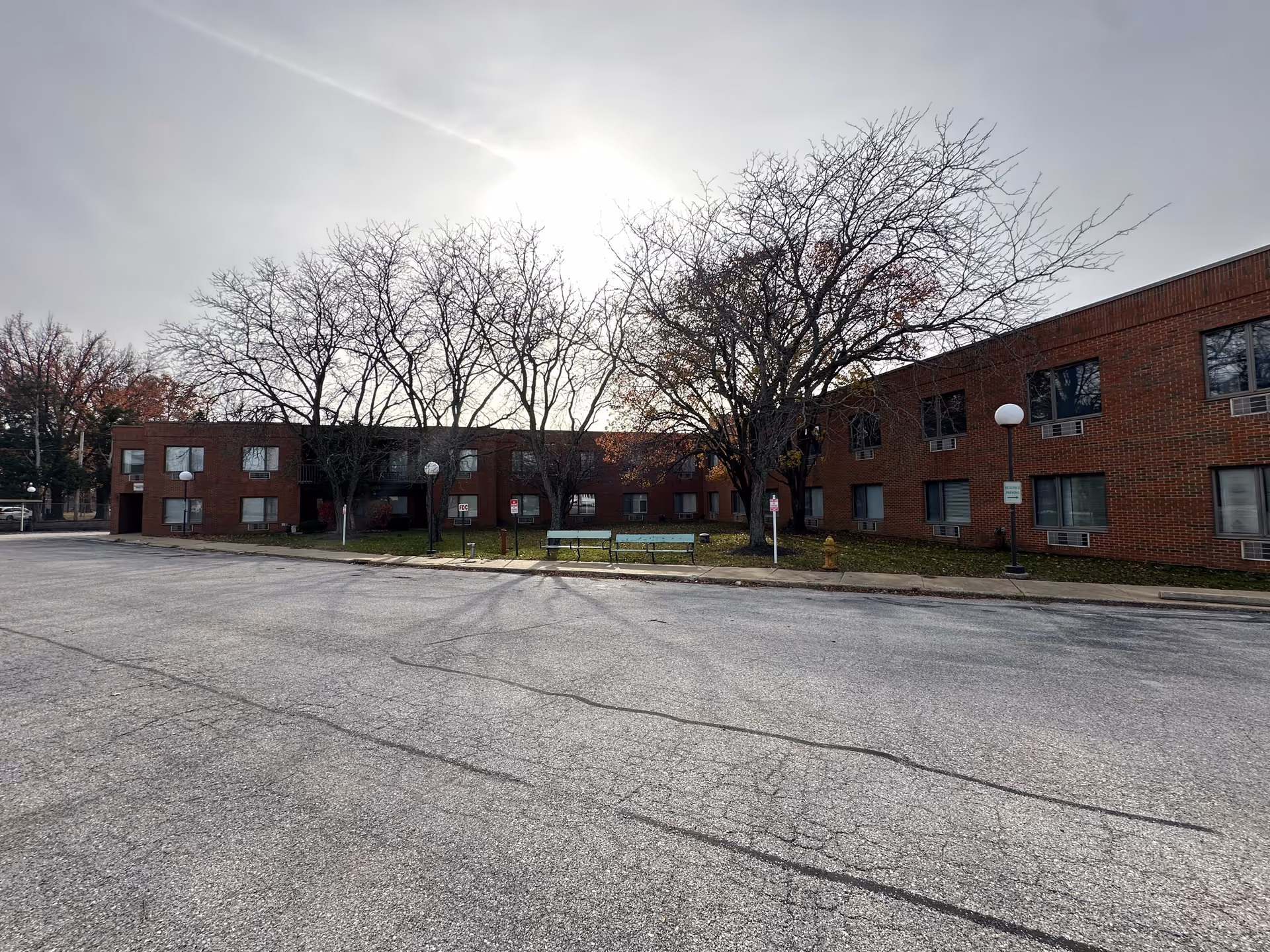 Two-story red-brick senior living building with bare trees, benches, lamp posts and an empty paved area in front under a bright overcast sky.