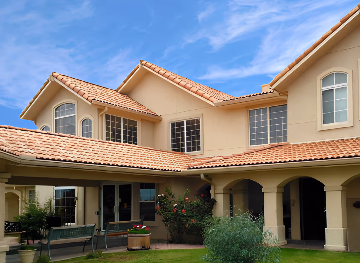 Exterior view of a senior living facility building with beige walls and terracotta roof tiles under a blue sky. The building features multiple windows and an arched covered walkway. There are benches and potted plants in the courtyard area with green grass and shrubs.
