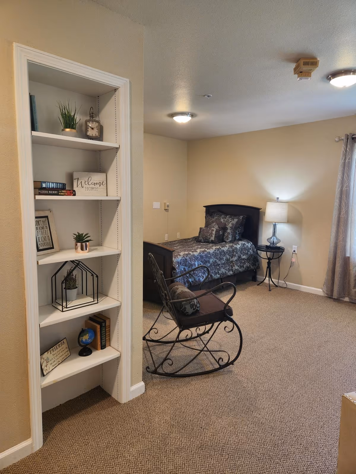 A cozy bedroom with beige walls and carpeted floor. The room features a single bed with patterned bedding and multiple pillows, a small round side table with a lamp, and a metal rocking chair with a cushion. To the left, there is a built-in white shelving unit decorated with books, small plants, a clock, and framed signs. A window with curtains is visible on the right side of the room.