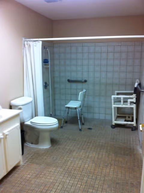 A spacious bathroom with beige tiled floor and walls. The room features a white toilet, a white vanity with a sink, a shower area with a white curtain, a shower chair, and grab bars for accessibility. There is also a white PVC pipe structure on wheels near the shower area.