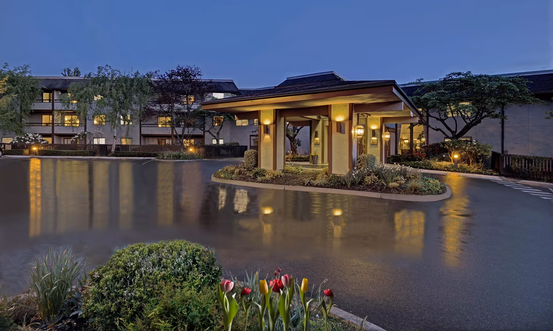 Covered porte-cochère entrance of a multi-story assisted living building at dusk with lit lanterns, wet driveway reflections, and flowers in the foreground.