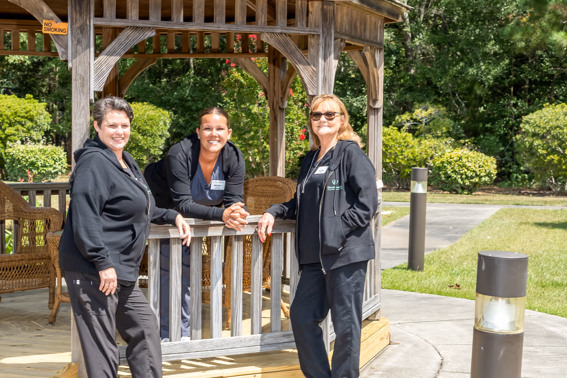 Three women wearing black jackets and name tags are smiling and posing together under a wooden gazebo in a garden area with green bushes and trees in the background. The gazebo has wicker chairs inside and a 'No Smoking' sign is visible on one of the wooden beams.