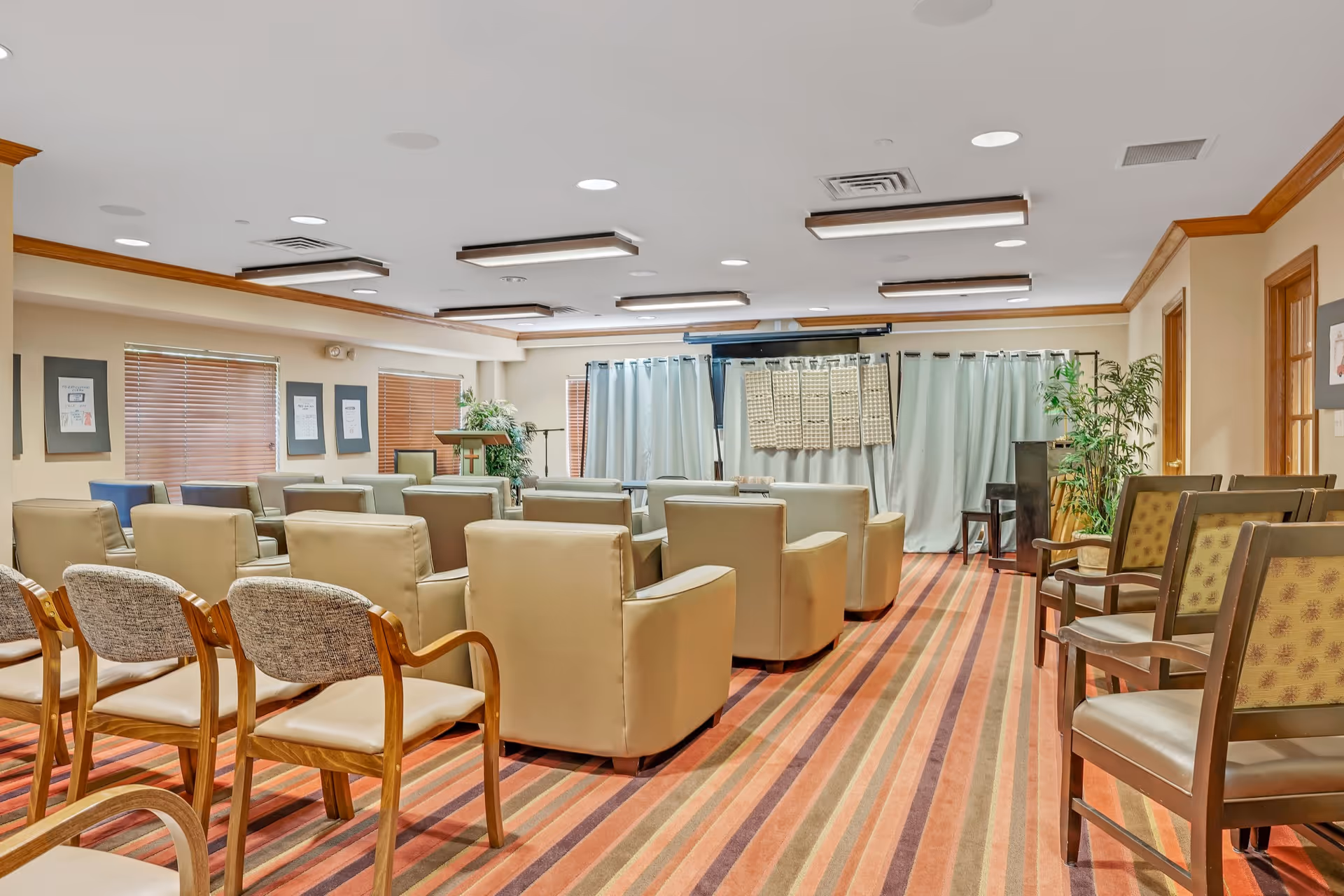 A spacious room with multiple rows of chairs and armchairs arranged facing a stage area with curtains and a podium, featuring a striped carpet and beige walls with wooden trim.