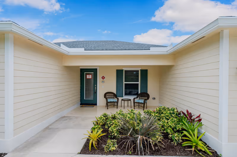 The entrance of The Caring Cottages facility featuring a door, two chairs, and landscaping.
