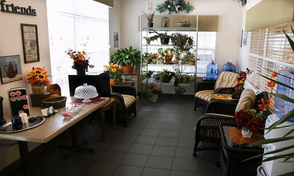 Sunlit communal seating area with wicker chairs, tables, many potted plants and water coolers.