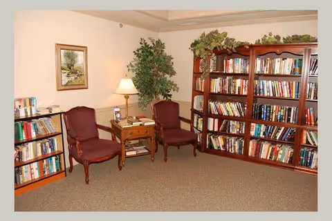 A cozy reading area with two upholstered armchairs in burgundy color, a wooden side table with a lamp, and two large wooden bookshelves filled with books. There is a framed painting on the wall and a potted plant in the corner, creating a warm and inviting atmosphere.