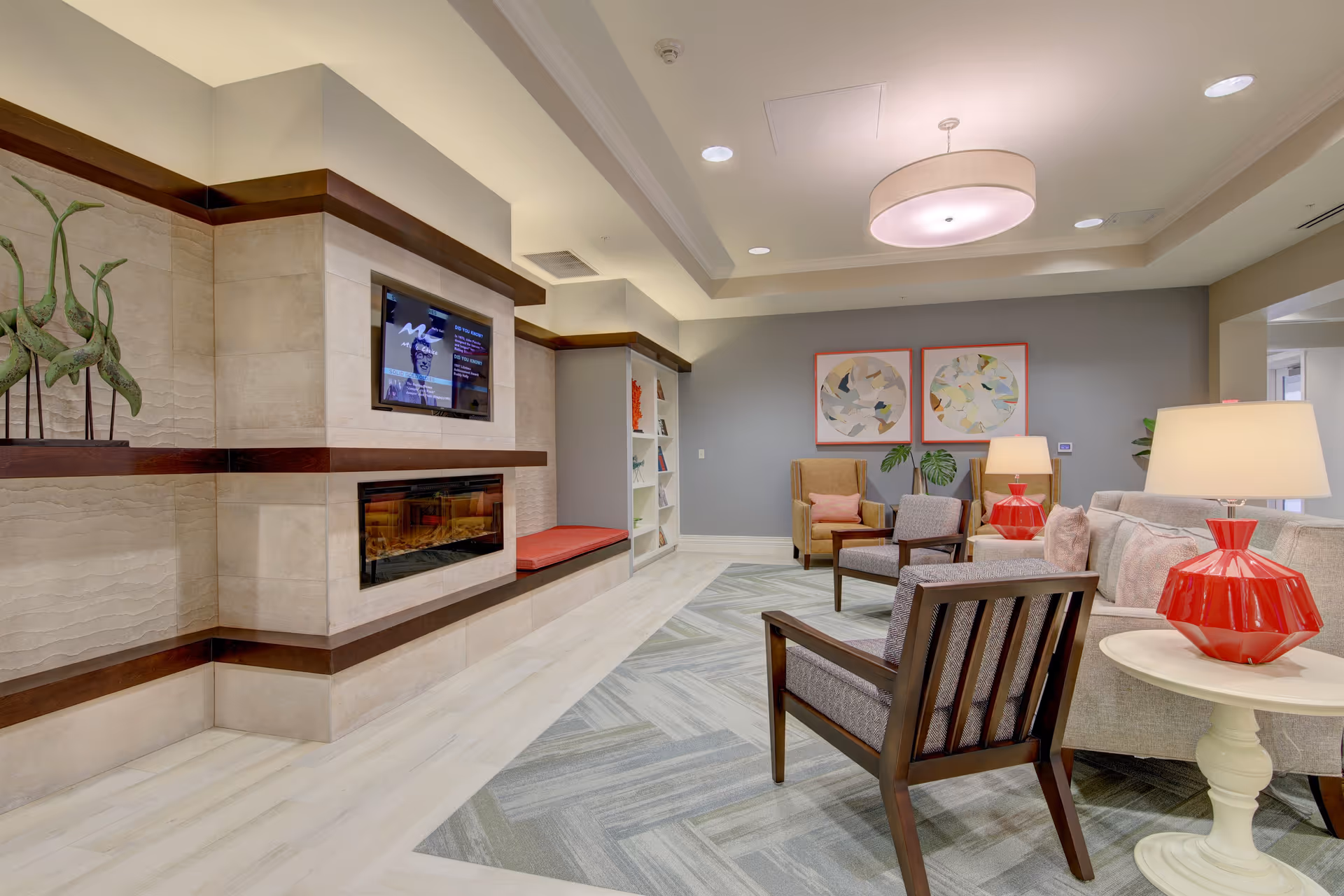 A modern and cozy living room area in an assisted living facility featuring a wall-mounted TV above a built-in fireplace, a cushioned bench, decorative sculptures, and a seating arrangement with armchairs, a sofa, side tables with red lamps, and abstract artwork on the wall.