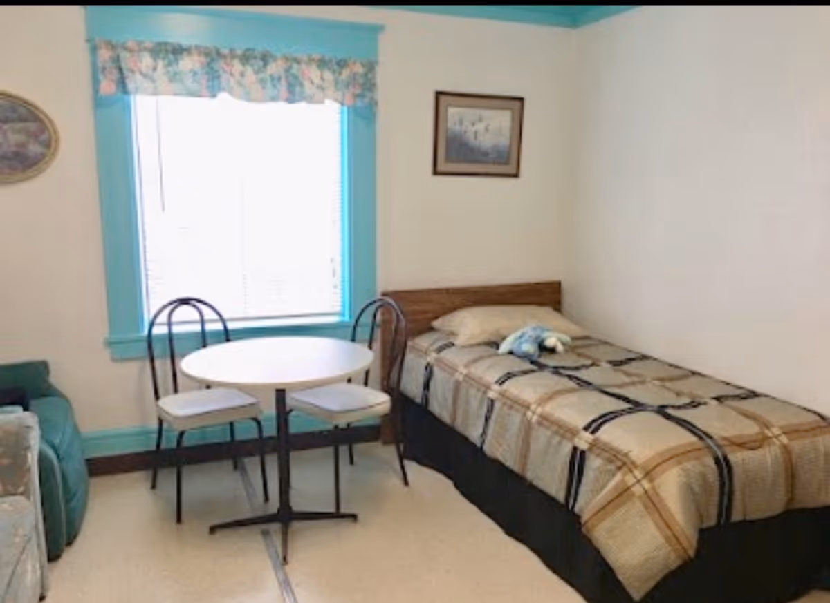 A small bedroom with a single bed covered in a plaid blanket and a pillow, a small round table with two chairs, a window with floral valance and turquoise trim, and a framed picture on the wall.