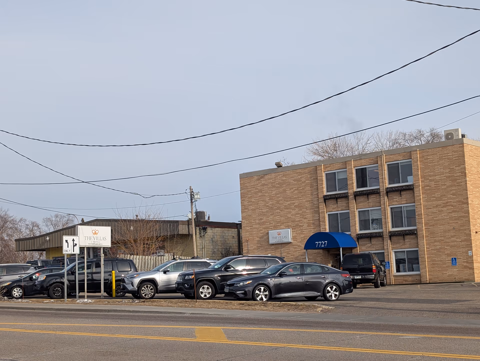 Front exterior of a brick senior living facility with a blue awning labeled '7727', a parking lot of cars, and a sign for The Villas.