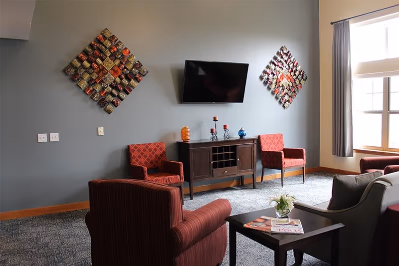 A cozy living room area with a wall-mounted flat-screen TV, two red patterned armchairs, a dark wooden cabinet with decorative items on top, and a small table with magazines and a flower vase. The room has a large window with gray curtains and two colorful square wall art pieces on a gray wall.