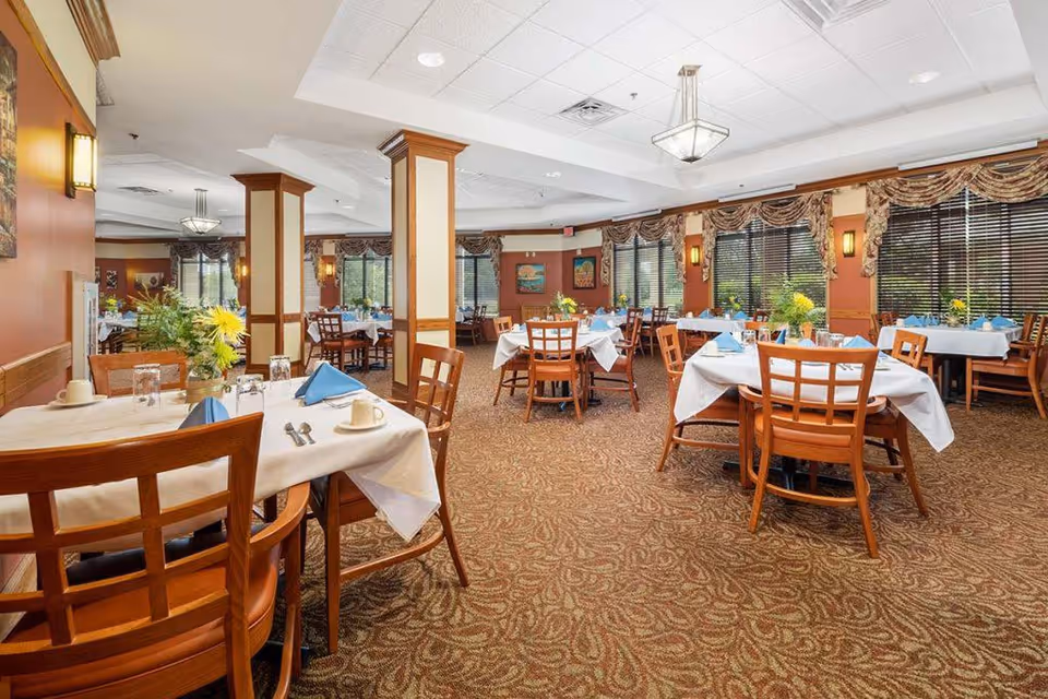 A spacious dining room with multiple tables covered in white tablecloths and set with blue napkins, cups, and silverware. The room features wooden chairs, patterned carpet, large windows with blinds and floral valances, and decorative wall sconces. There are columns in the center of the room and framed artwork on the walls.