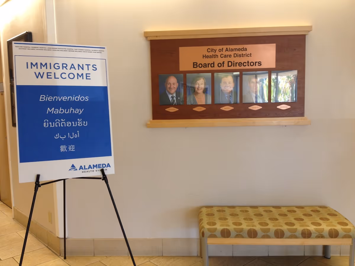 Interior hallway with a sign on a stand that reads 'IMMIGRANTS WELCOME' in multiple languages and a wooden board on the wall displaying photos and names of the City of Alameda Health Care District Board of Directors. A patterned bench is placed below the board.