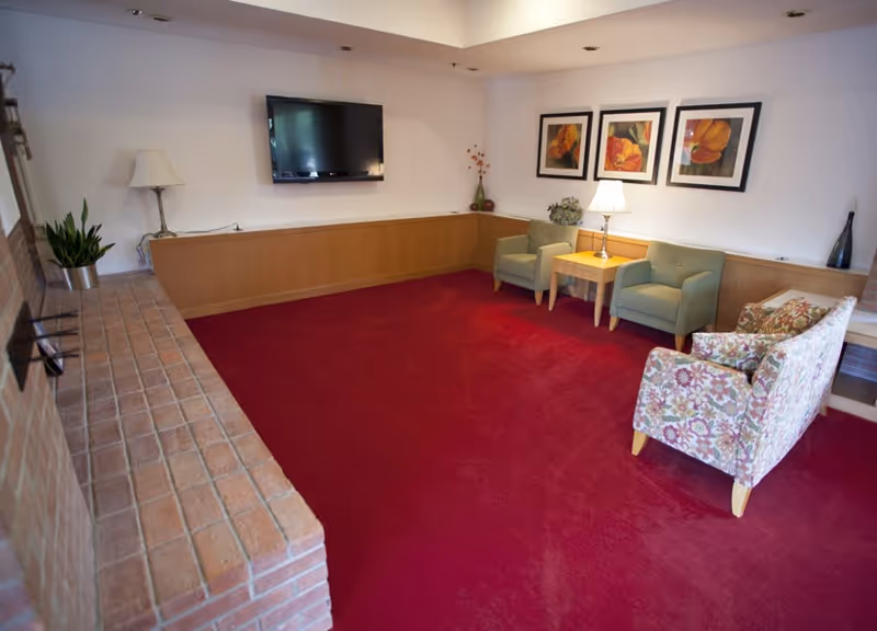 A cozy living room with a red carpet, two green armchairs, a floral-patterned loveseat, a wooden side table with a lamp, three framed floral artworks on the wall, a flat-screen TV mounted on the wall, and a brick ledge along one side.