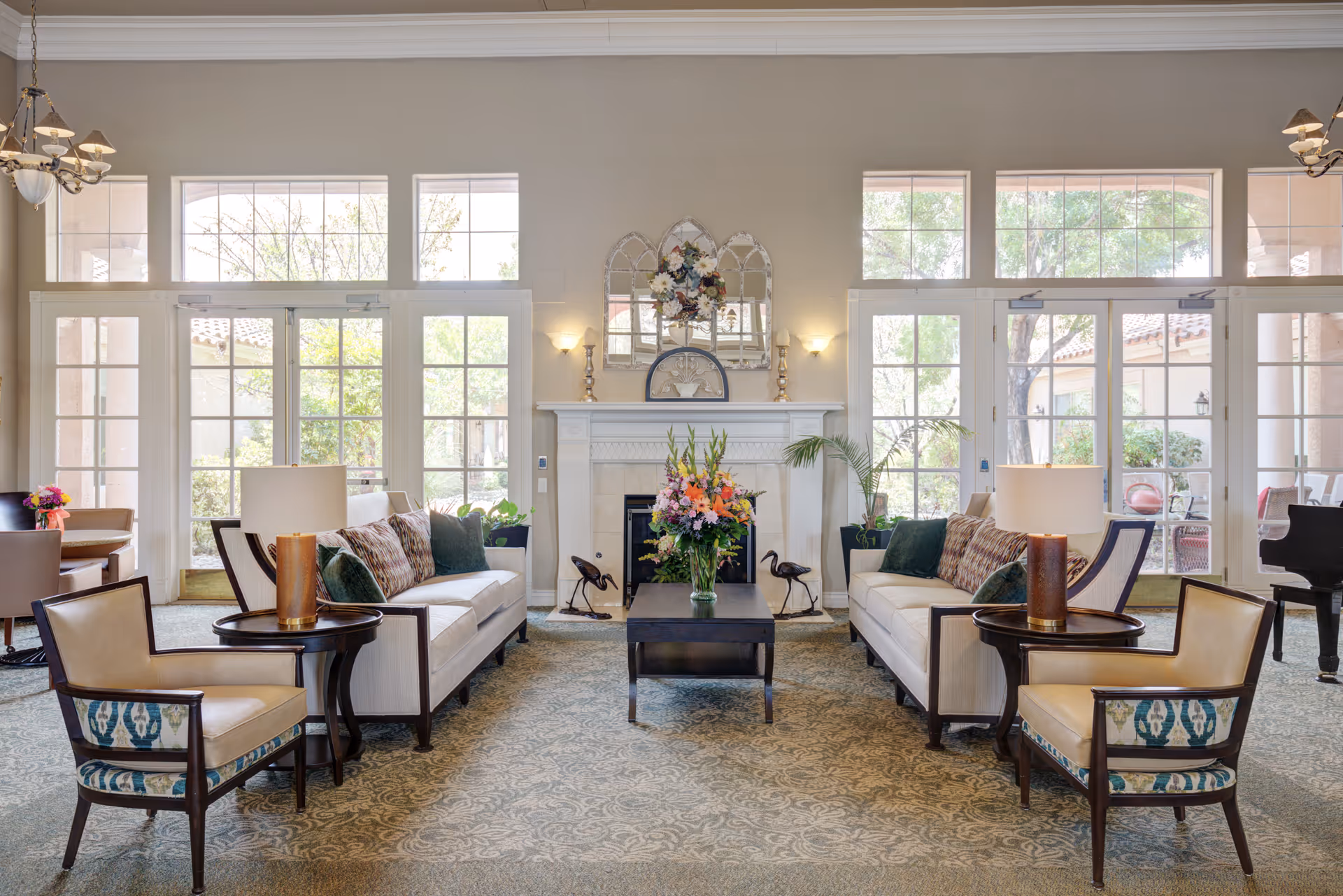 Spacious, well-lit communal living room with two sofas and armchairs arranged around a coffee table and fireplace, with large windows and glass doors.