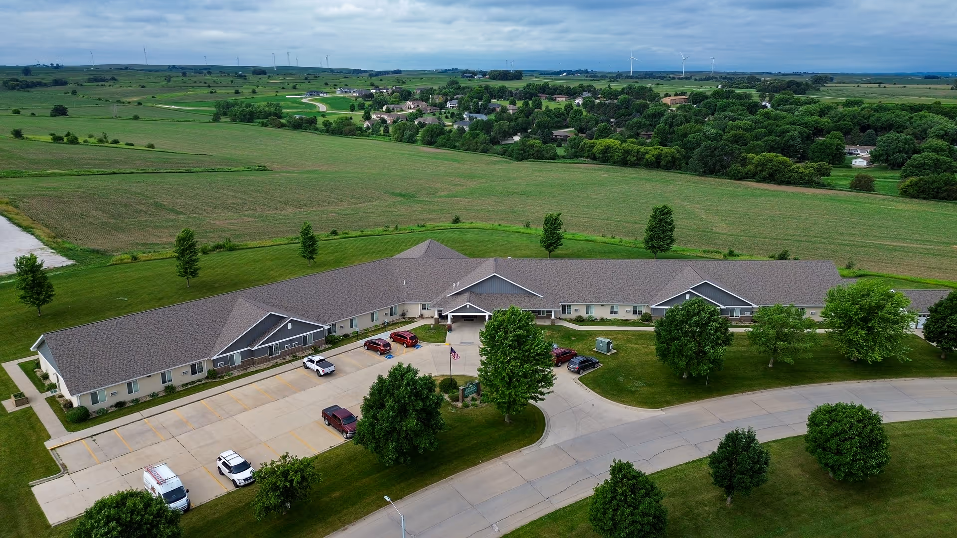 Aerial view of a single-story senior living facility named Golden Horizons of Ida Grove surrounded by green lawns and trees, with a parking lot in front and open fields in the background under a cloudy sky.
