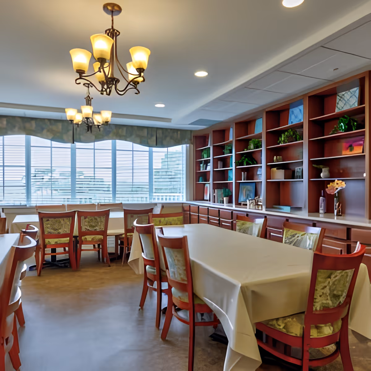 A dining room with tables covered in white tablecloths and wooden chairs with patterned cushions. The room features large windows with blinds, a built-in wooden shelving unit with decorative items and plants, and two ornate chandeliers hanging from the ceiling.