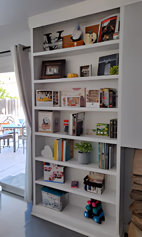 A white built-in bookshelf filled with various decorative items including framed pictures, books, small plants, and colorful art pieces. To the left, there is a sliding glass door with a curtain, leading to an outdoor patio area with a table and chairs.
