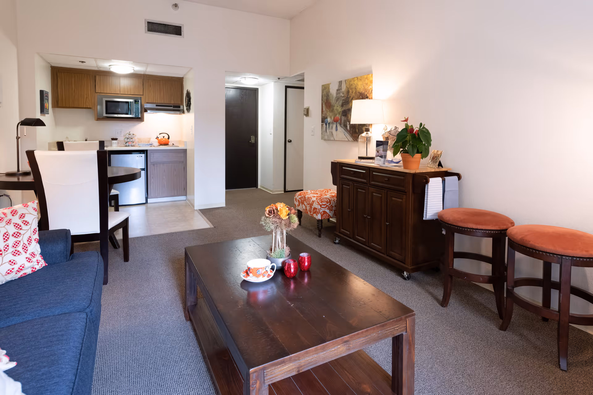 Interior view of a senior living facility apartment featuring a living room with a blue sofa, a dark wooden coffee table with a cup and decorative items, two orange cushioned stools, a wooden sideboard with a lamp and plant, and a small kitchen area with wooden cabinets, a microwave, and a mini fridge in the background.