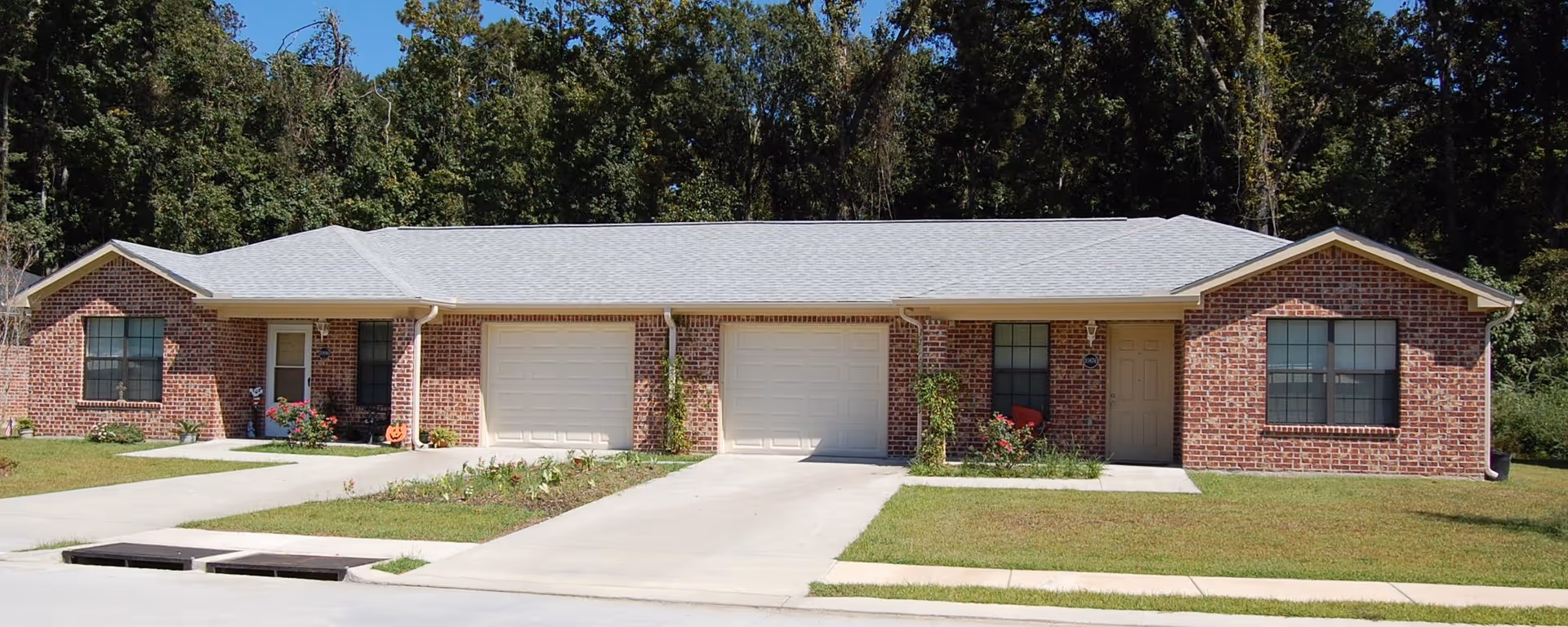 Exterior view of a single-story brick residential building with two garages and two front doors, surrounded by grass and trees in the background.