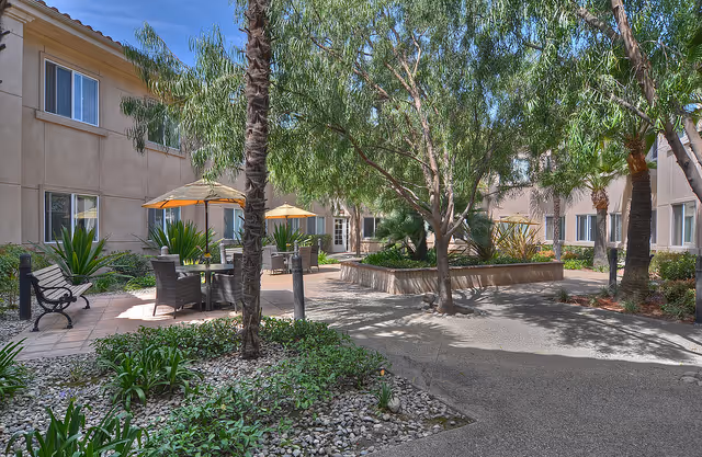 Shaded courtyard with patio tables and umbrellas, benches, trees, and a surrounding two-story building.