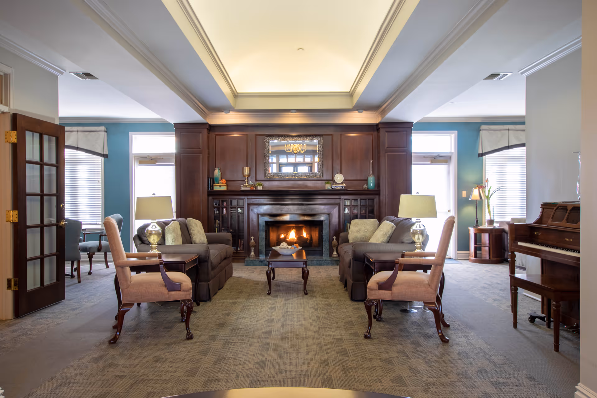 A cozy living room with two sofas facing each other and a wooden coffee table in between. There are two armchairs on either side of the sofas. The room features a lit fireplace with a wooden mantel and a decorative mirror above it. The walls are painted light blue and white, and there is a piano on the right side of the room. The room is well-lit with natural light coming through windows with blinds and valances.