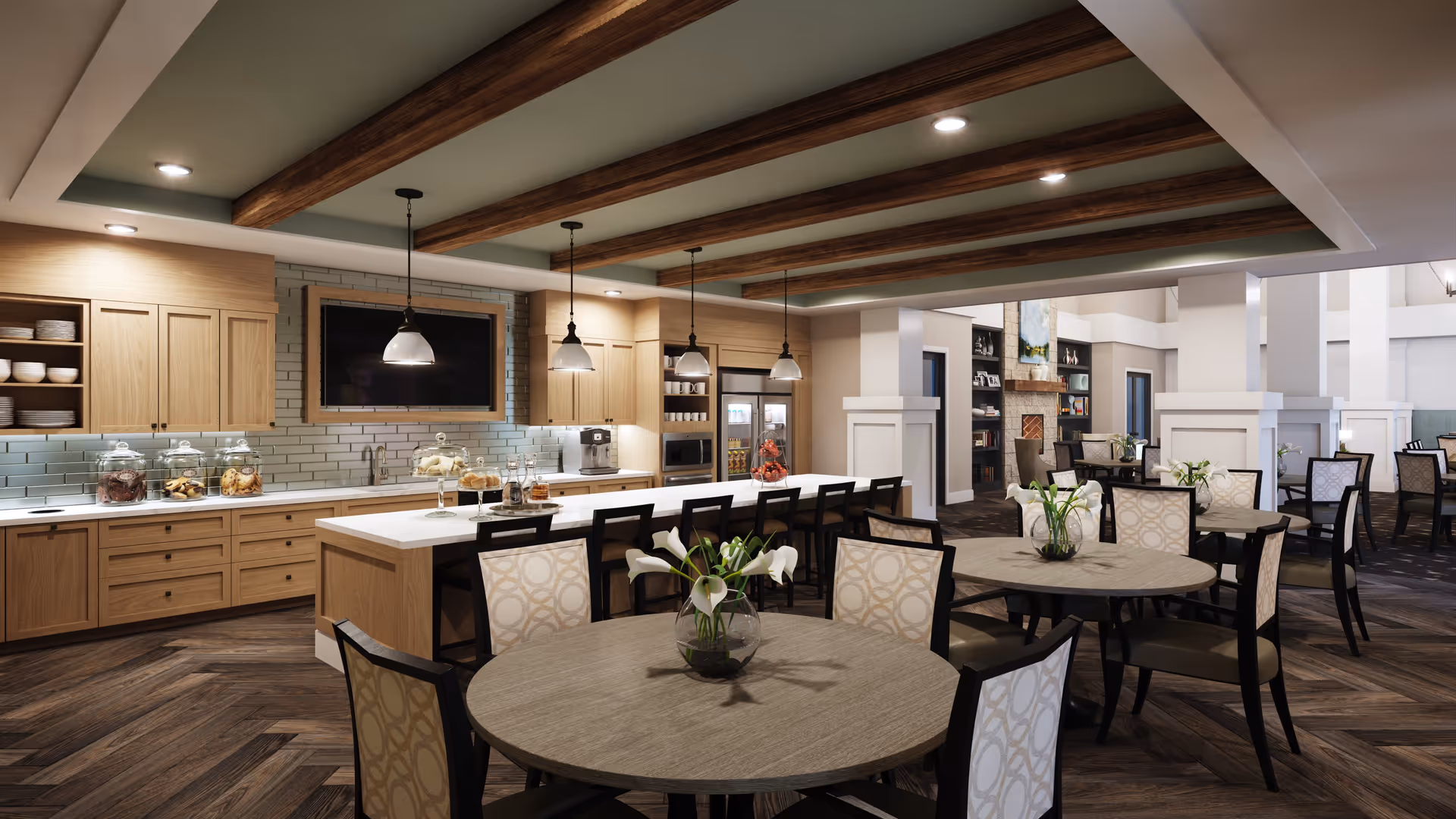 A spacious and modern dining area with round tables and cushioned chairs, each table decorated with a vase of white flowers. The room features a kitchen area with wooden cabinets, a long island with bar stools, pendant lights hanging from a ceiling with wooden beams, and a wall-mounted TV. The background shows a cozy lounge area with a fireplace and bookshelves.
