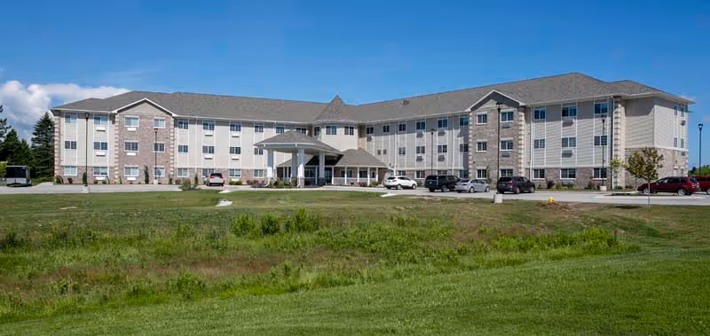 Front view of a three-story senior living facility with a covered entrance, parked cars, and a large grassy lawn under a blue sky.