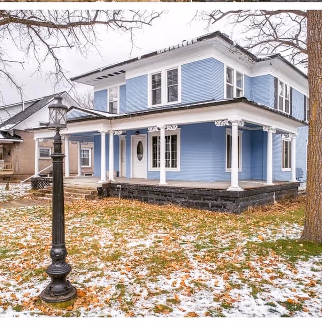 Exterior view of a two-story blue house with white trim and a wrap-around porch supported by white columns. The yard has patches of snow and fallen leaves, and there is a black vintage-style street lamp in the foreground. Trees with bare branches surround the house.