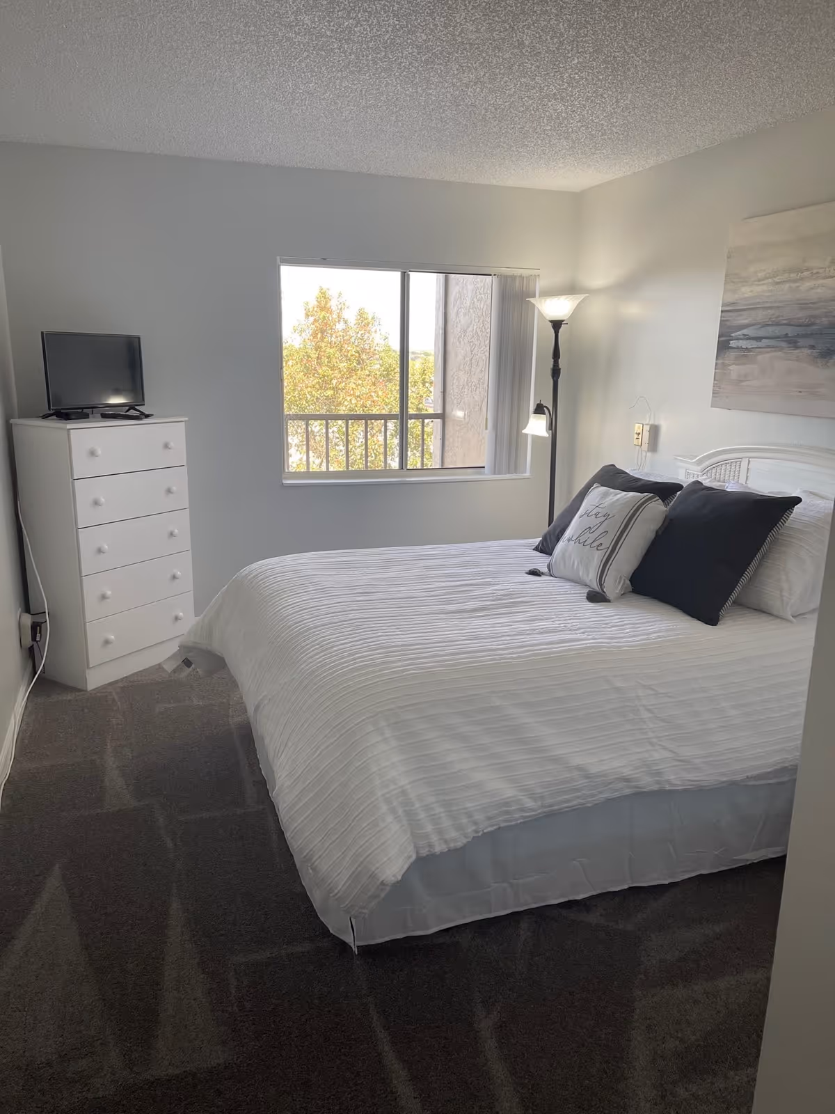 A bright bedroom with a white bedspread and three pillows, including one with the text 'stay awhile'. There is a white dresser with a small TV on top, a window showing trees outside, a floor lamp with two lights, and a piece of abstract wall art above the bed.