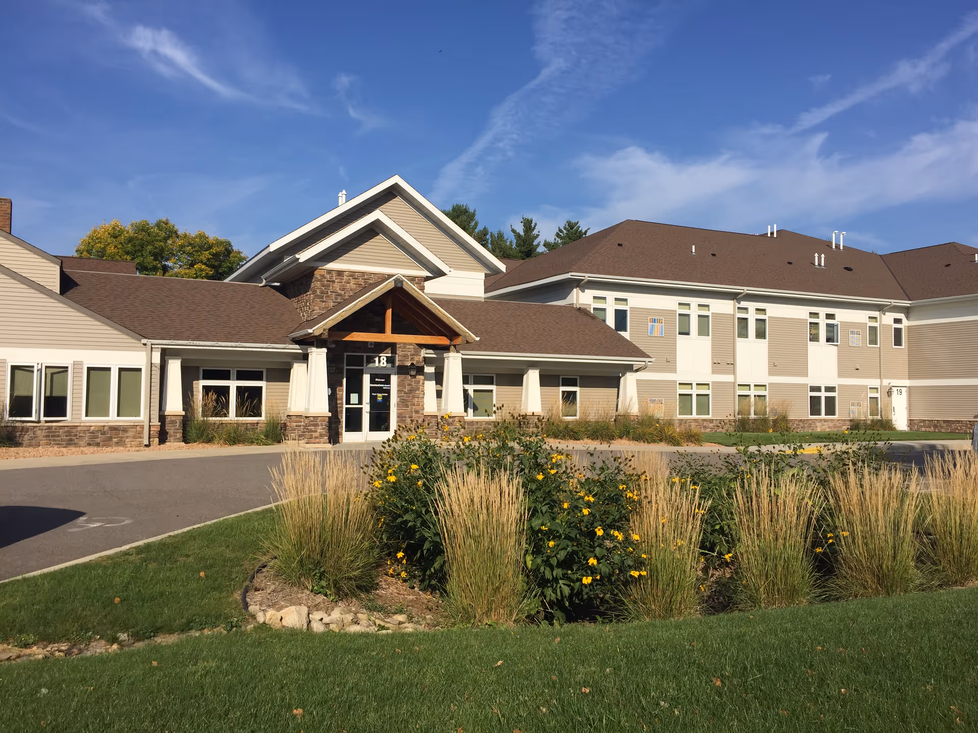 Exterior view of Melrose Park View Place, a two-story senior living facility with beige siding and stone accents. The building has a peaked roof and a covered entrance with the number 18 above the door. In front of the building is a landscaped area with tall grasses and yellow flowers, and a paved driveway leading to the entrance. The sky is clear with some wispy clouds.