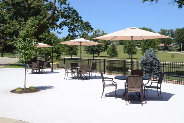 Outdoor patio area with several round tables and chairs, each shaded by large beige umbrellas. The patio is surrounded by a black metal fence and overlooks a green grassy area with trees under a clear blue sky.