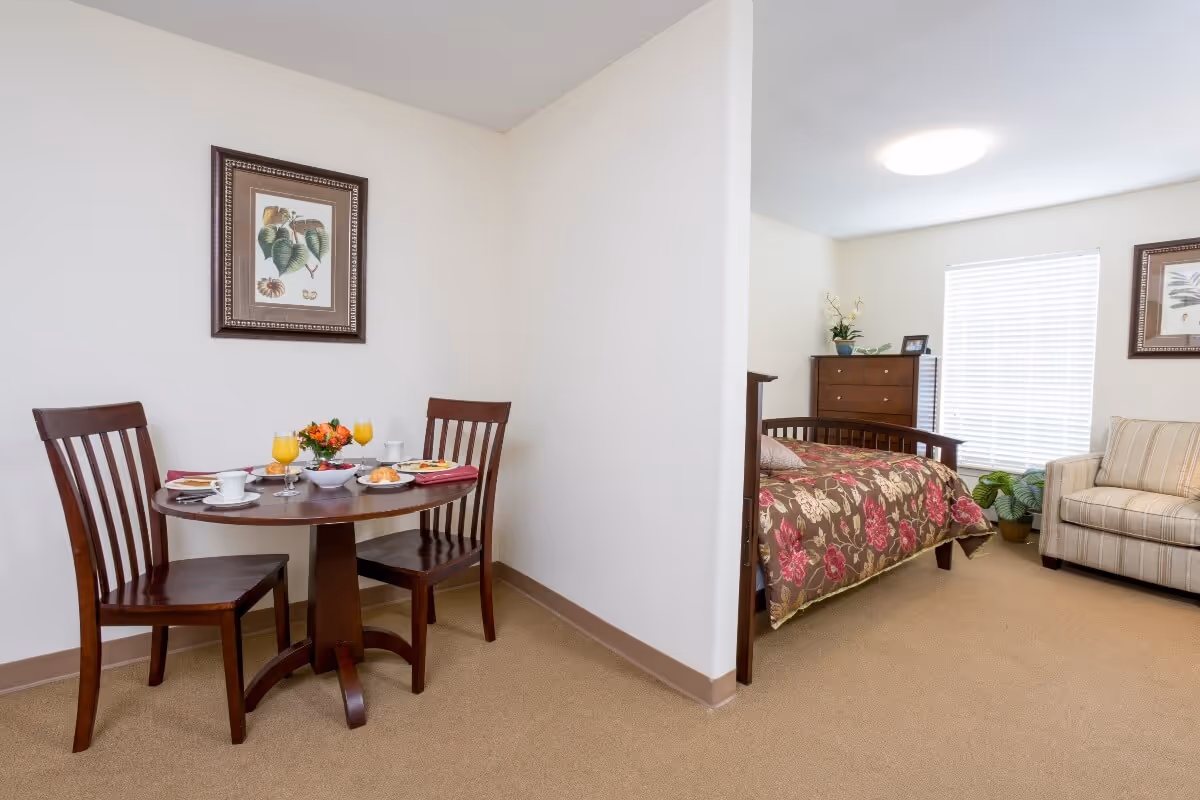 A bright senior living studio with a round dining table set for two and a bed and sofa visible in the background.