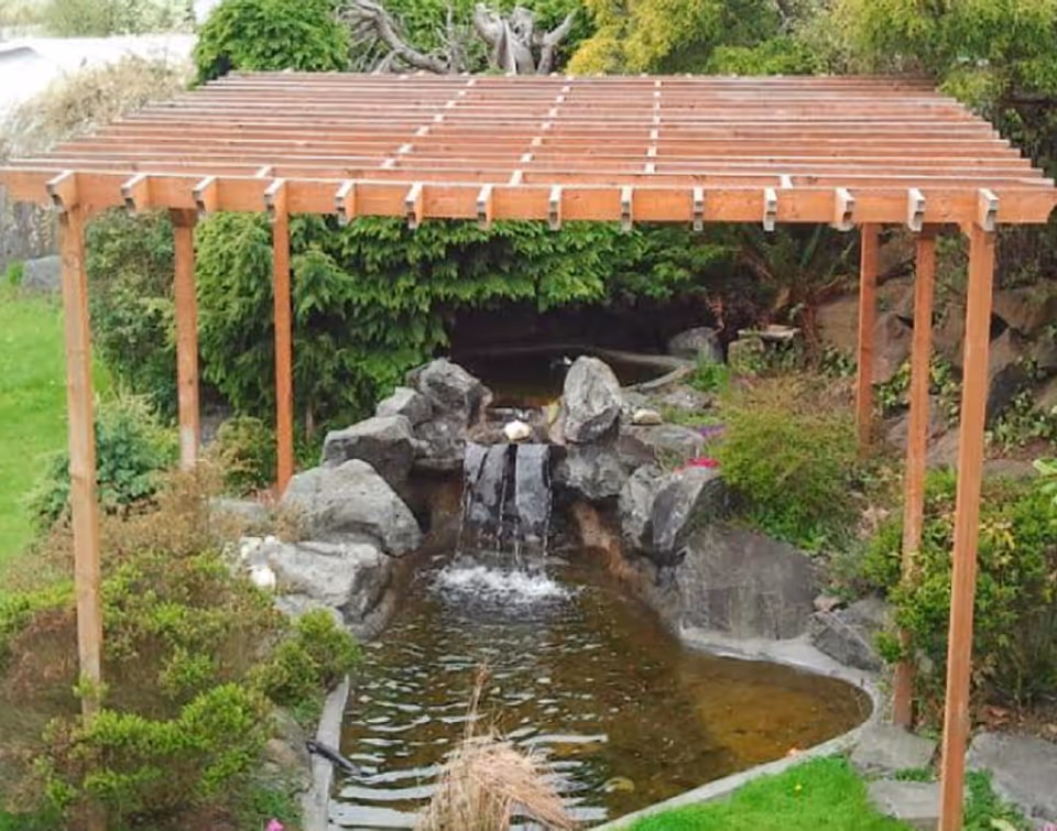 Wooden pergola over a landscaped pond with rocks and a small waterfall surrounded by greenery.