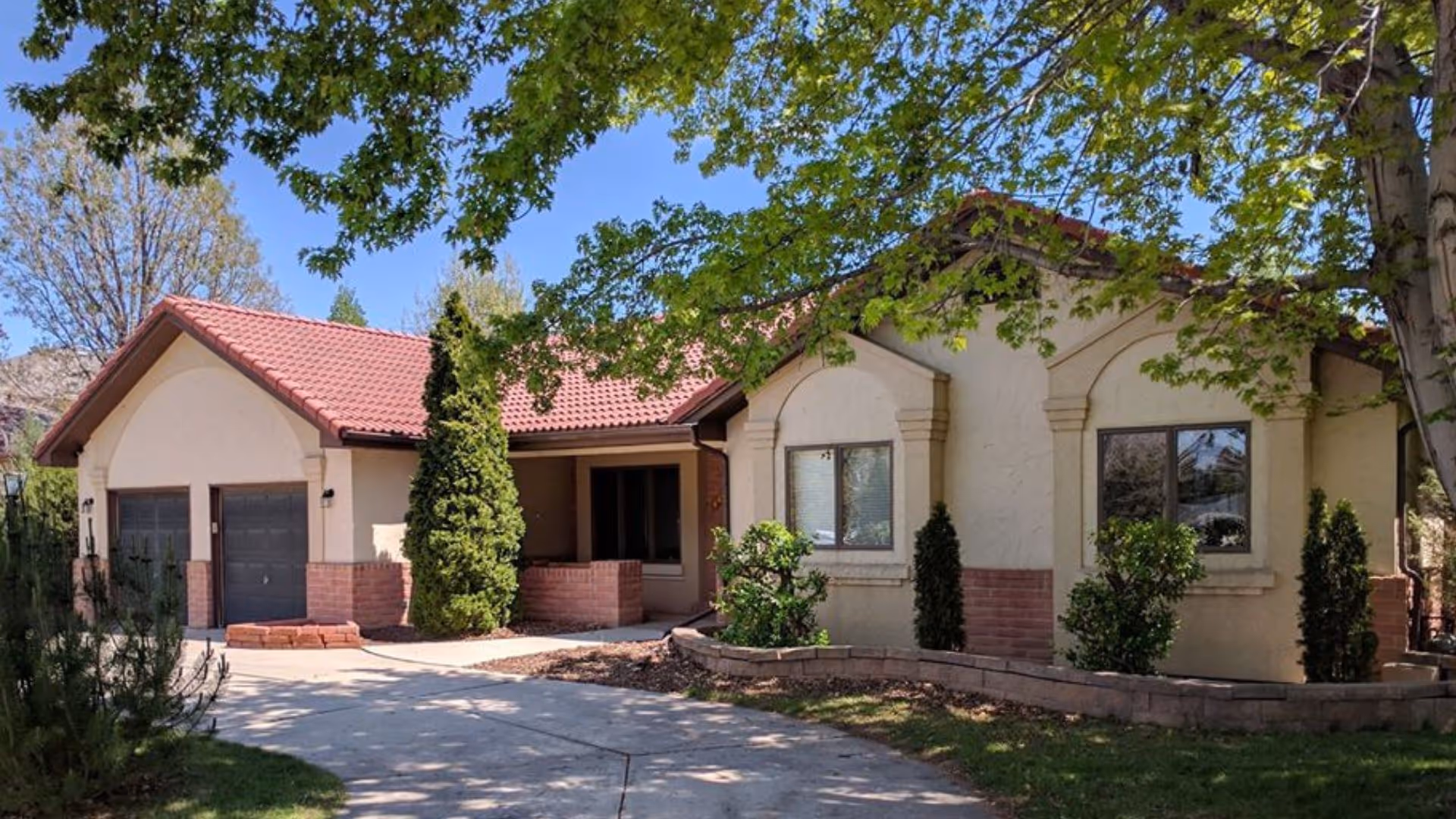 Exterior view of a single-story residential building with a red tiled roof, beige walls, and a two-car garage. The driveway curves in front of the house, and there are several green trees and shrubs around the property under a clear blue sky.