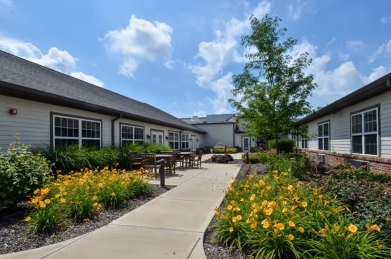 Outdoor courtyard between two single-story building wings with a paved walkway, seating, yellow flowers, and a tree under a blue sky.