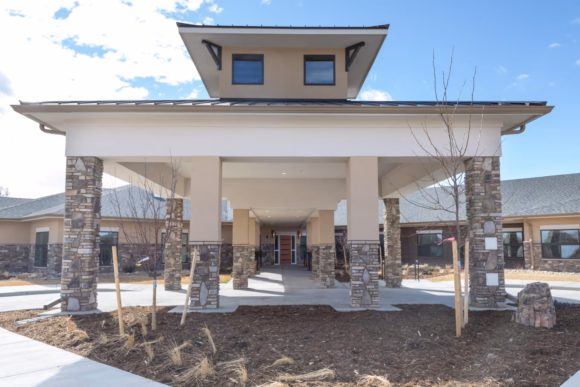 Front exterior view of a senior living facility named Accel At Longmont, featuring a covered entrance supported by stone and beige pillars, with a clear blue sky in the background.