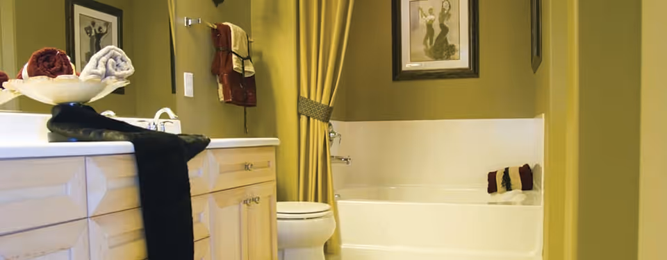 A bathroom with a white bathtub and shower combination, a toilet, and a vanity with a sink. The vanity has light wood cabinets and a white countertop with rolled towels placed on it. The walls are painted olive green, and there is a framed picture hanging above the bathtub. A yellow shower curtain is partially drawn to the side.