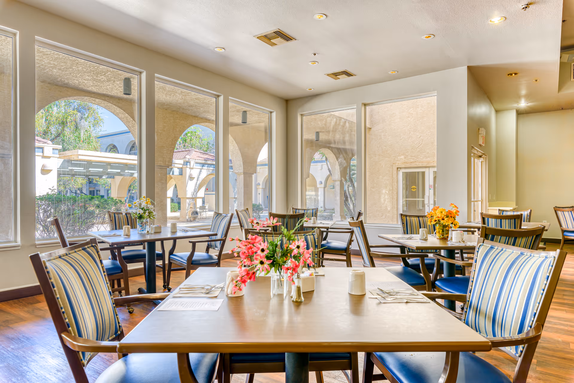 Bright dining room with multiple tables and chairs, each table decorated with flower arrangements. Large windows provide a view of an outdoor courtyard with arches and greenery.