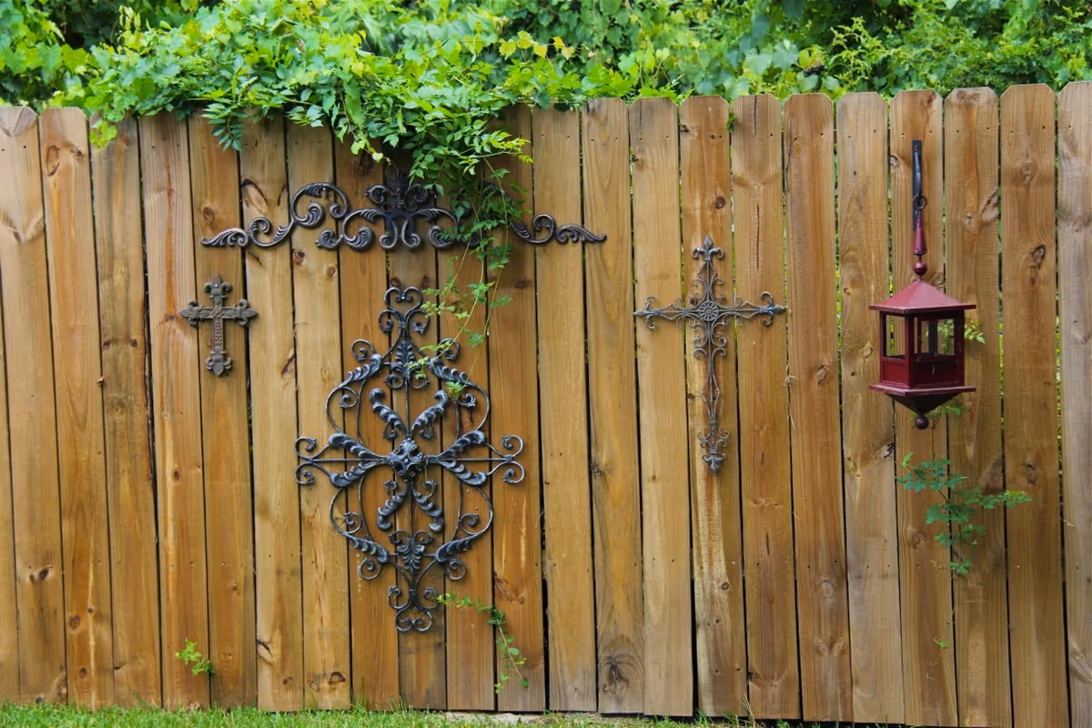 A wooden fence with three decorative metal crosses and a red bird feeder hanging on it. Green foliage is visible at the top and some grass at the bottom.