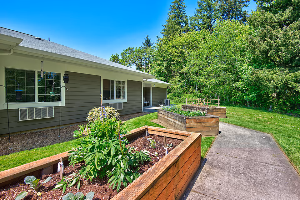 Outdoor garden area at Parkland Village Retirement Community featuring raised wooden garden beds with plants and flowers, a concrete pathway, and a building with windows and a covered porch. The background includes lush green trees under a clear blue sky.