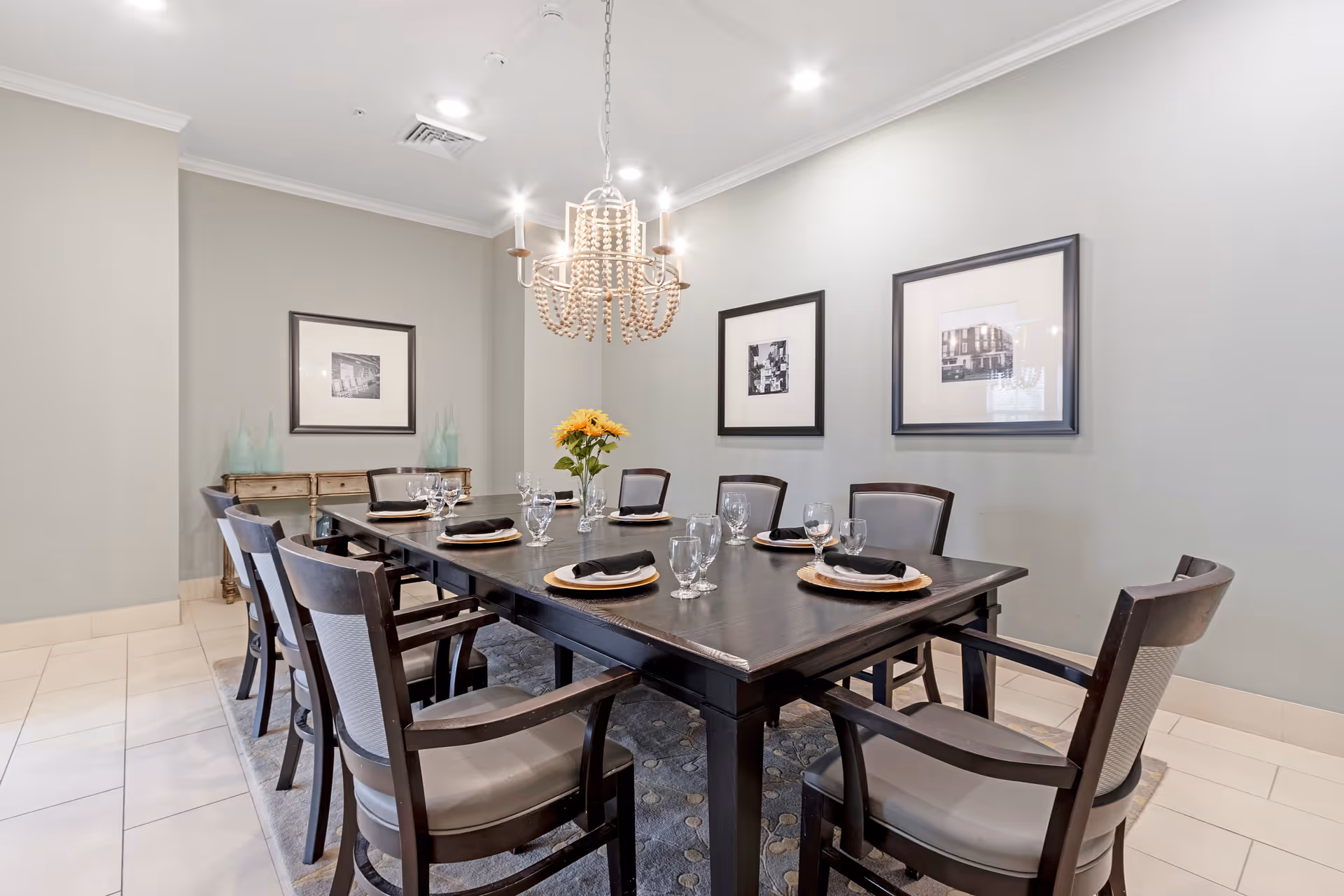 A dining room with a dark wooden rectangular table set for eight people. Each place setting includes a plate with a black napkin and glassware. The room features light gray walls with three framed black and white photographs, a chandelier with a beaded design hanging above the table, and a sideboard with decorative glass vases. The floor is tiled with a light-colored rug under the table.