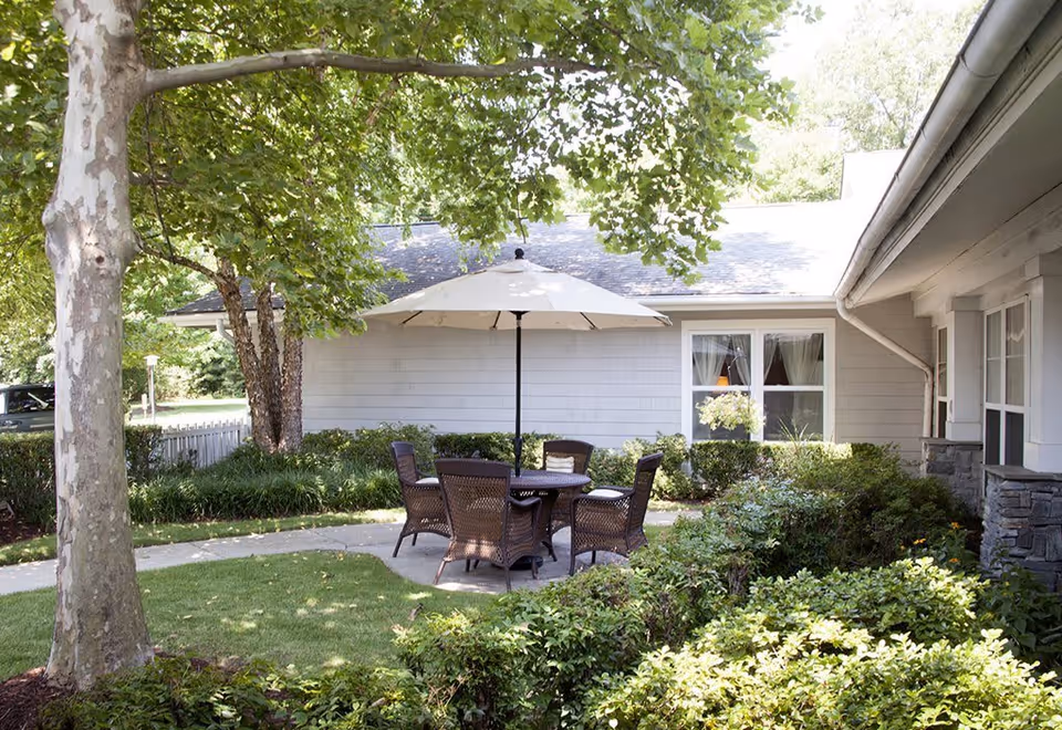 Outdoor patio area at Sunrise of Richmond featuring a round table with five wicker chairs and a large white umbrella. The patio is surrounded by green bushes, trees, and a well-maintained lawn, with a light gray building in the background.