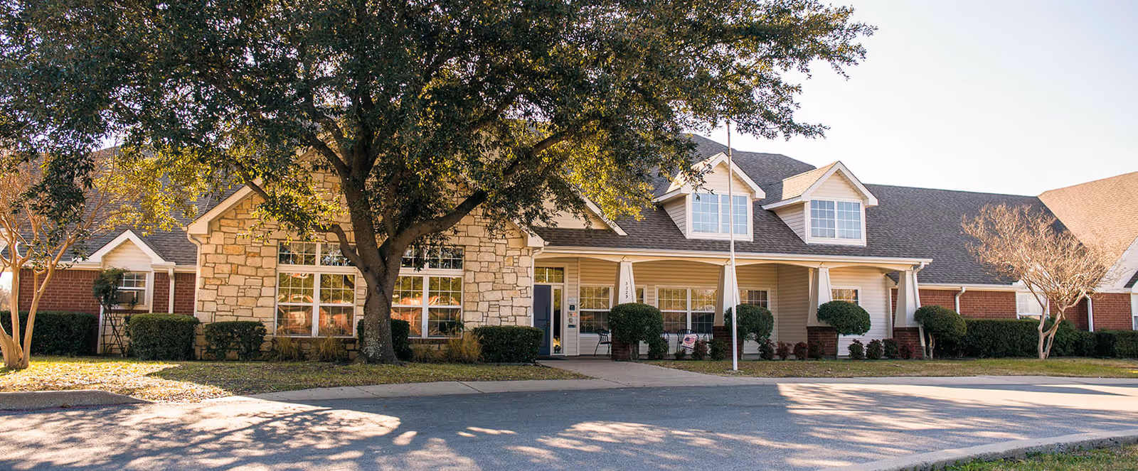Exterior view of a single-story senior living facility building with a stone and brick facade, large windows, a covered porch with white columns, and a large tree in front casting shadows on the driveway.